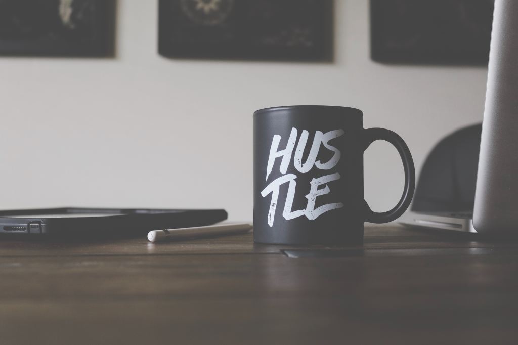 A black coffee mug with the word 'Hustle' printed on it, sitting on a desk by a laptop and a pencil.