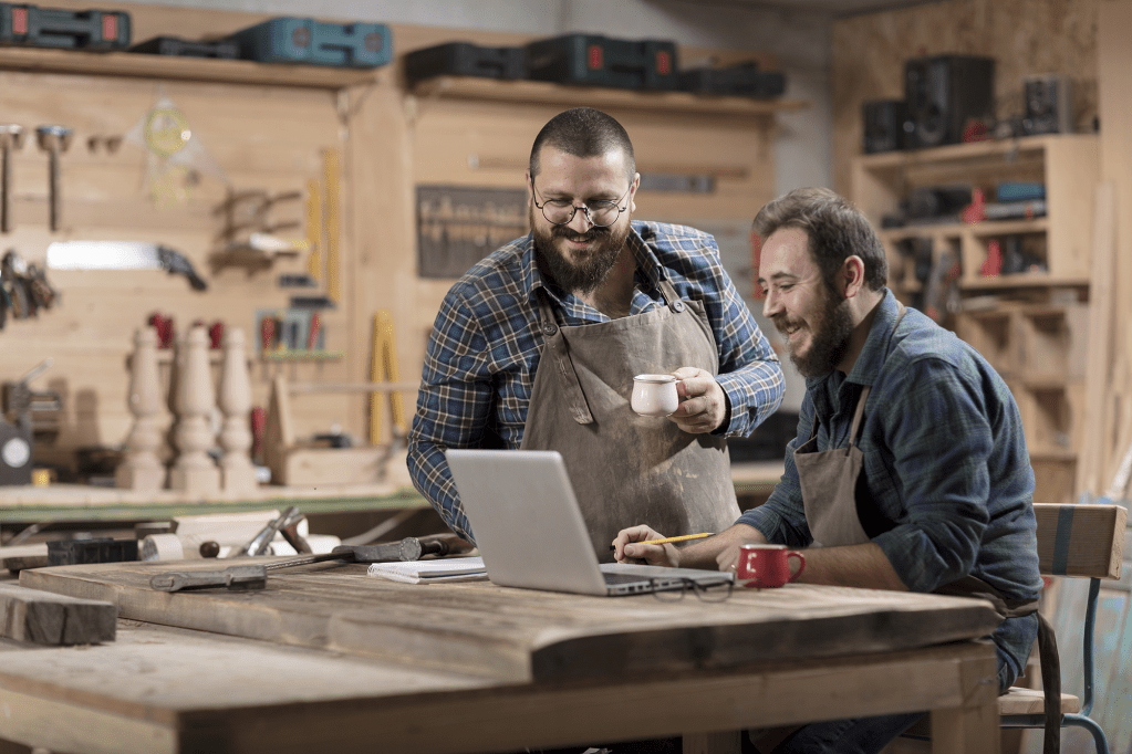 Two men in a woodworking shop calculate their overhead costs on a laptop