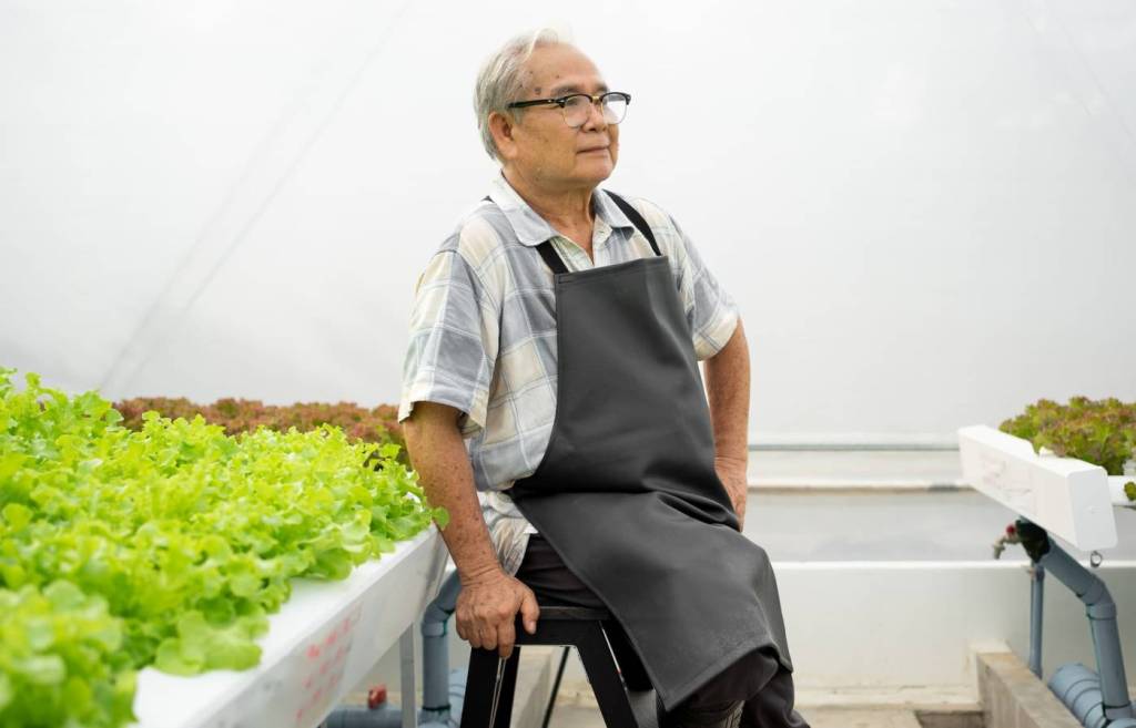 An older Asian man in an apron sits against a styrofoam planter will with green vegetables considering the cost of small business insurance