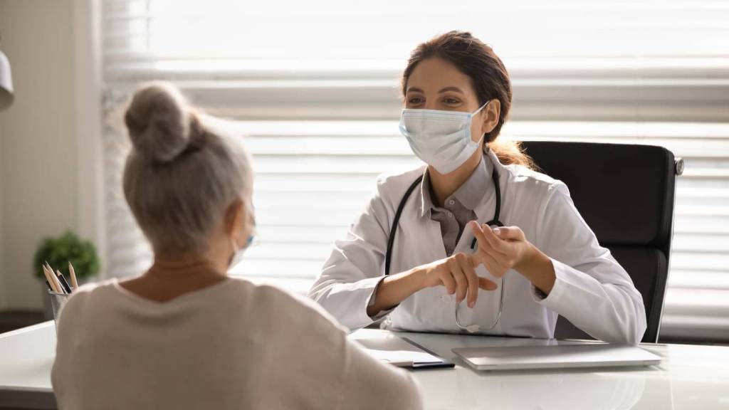 A young doctor in a mask sits across the desk from a patient